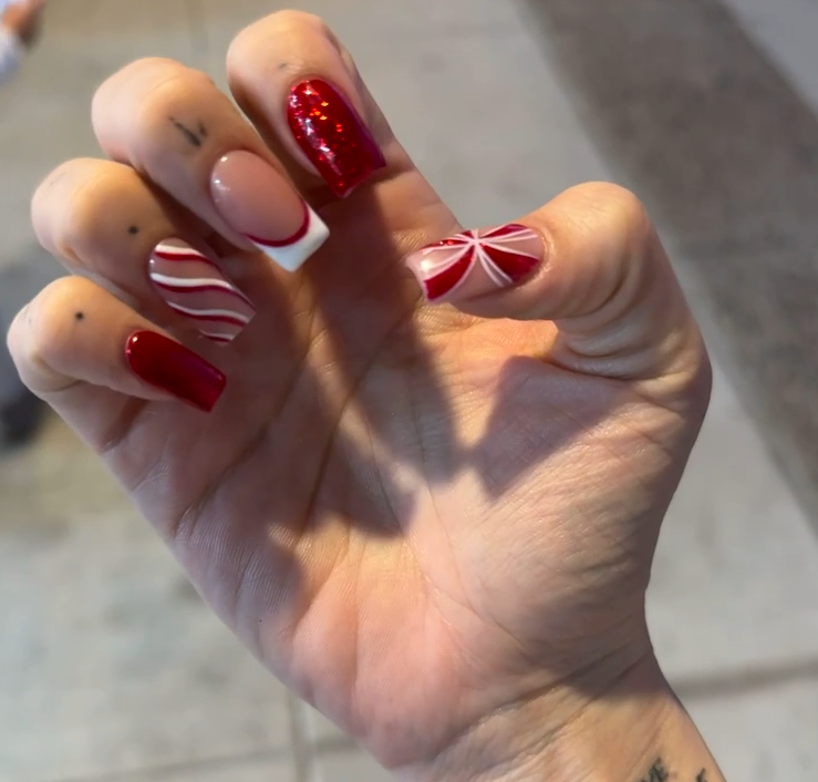 Close-up of a hand showcasing Christmas-themed nail art with red and white designs, including glossy red, candy cane stripes, and a bow motif