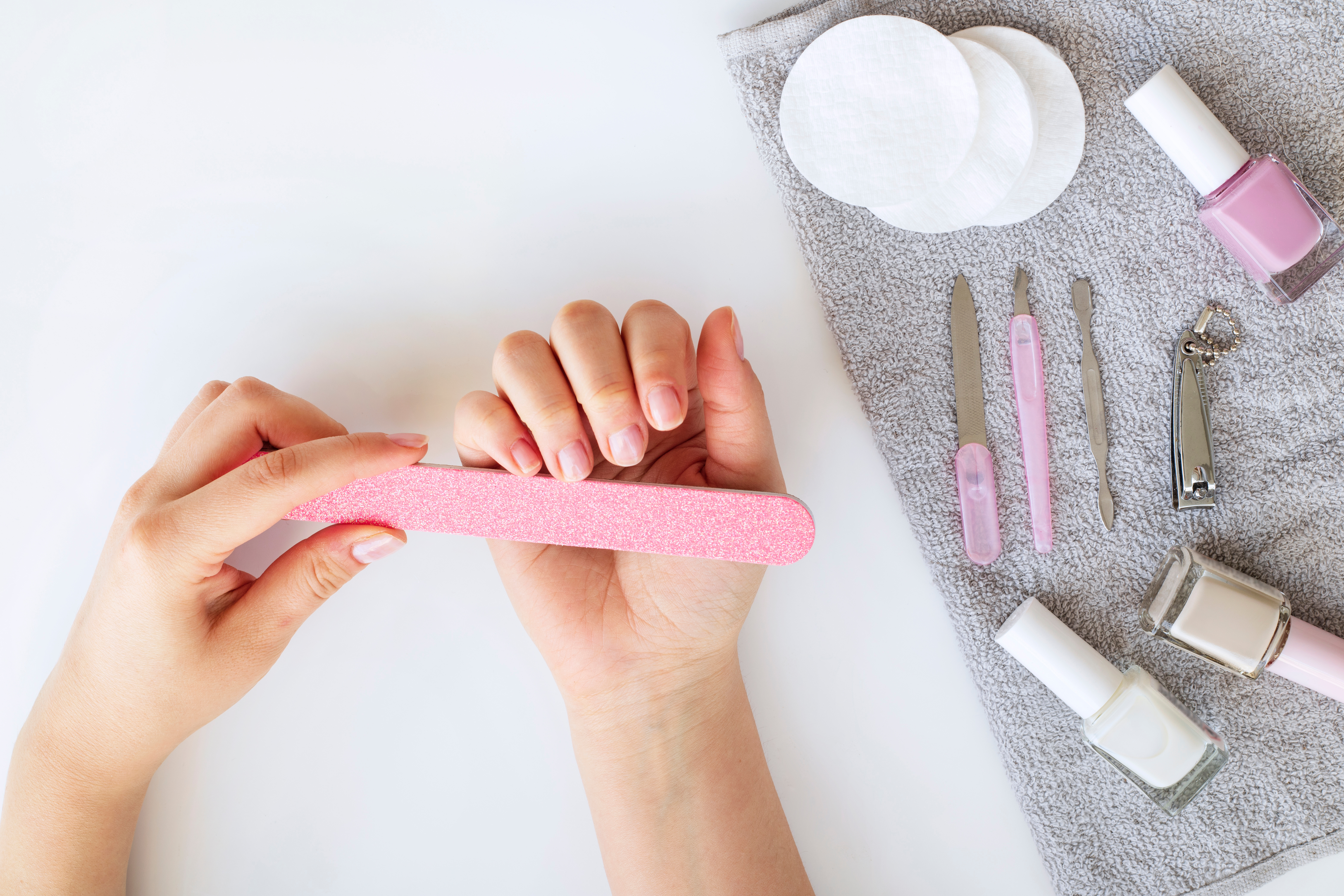 Person using a pink nail file with manicure tools and nail polish bottles on a gray towel