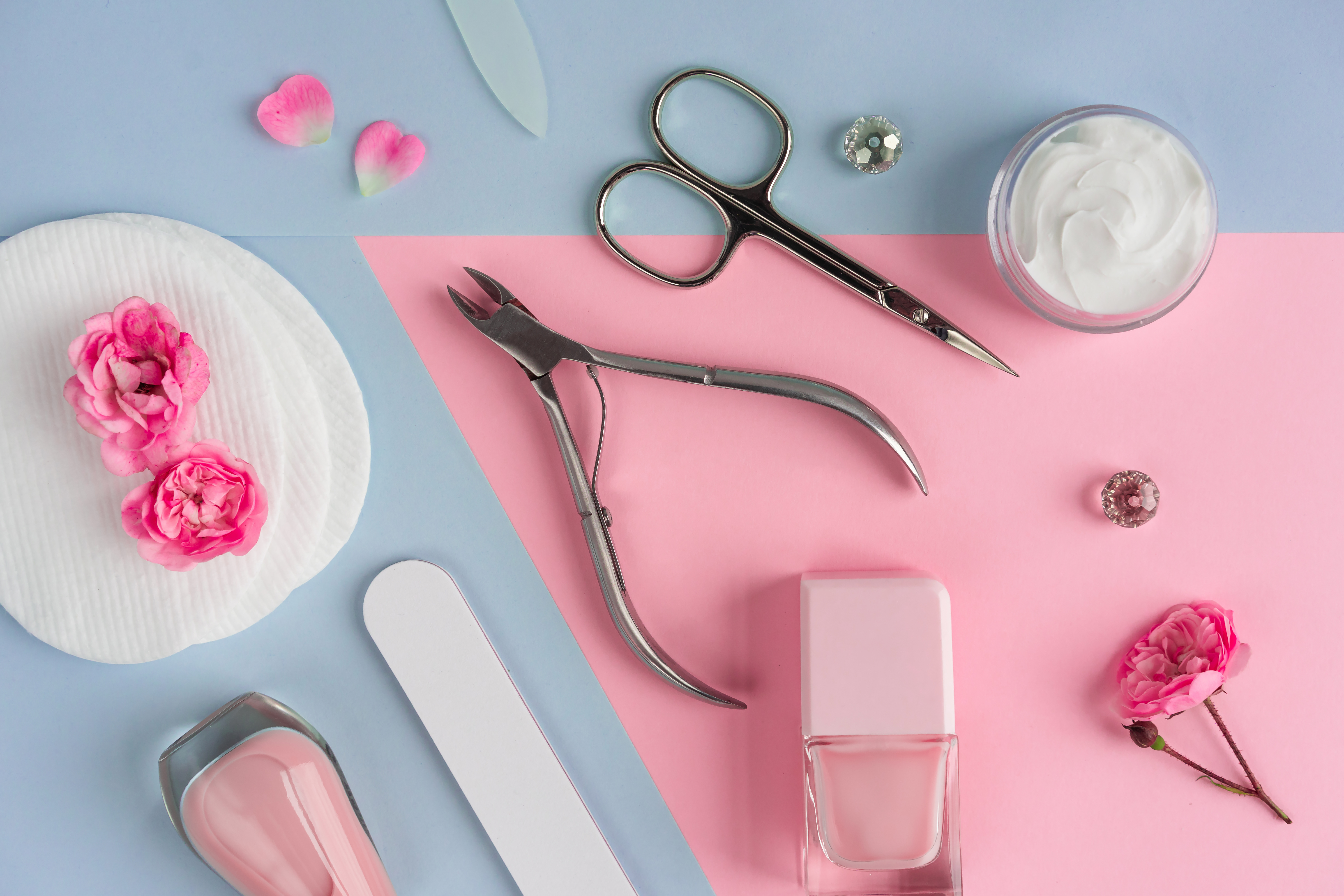 Nail care accessories with nail polish, scissors, cuticle nipper, nail file, and beauty cream on a dual-tone pink and blue background